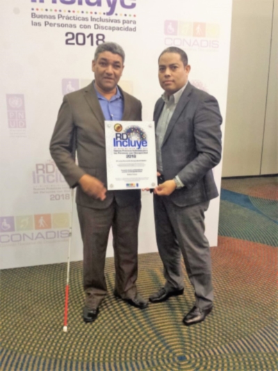 El presidente de la Federación Dominicana de Ciegos, Lino Delgado, posa con el premio junto al ex coordinador del Programa ÁGORA República Dominicana, Miguel González